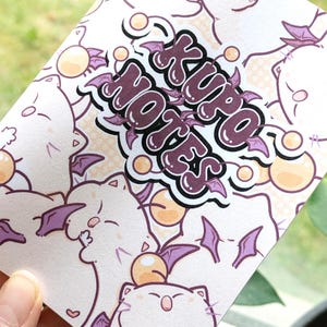 May include: A white notebook with a repeating pattern of cartoon cat-like creatures with purple bat wings and orange orbs. The words "Xuro Notes" are in a purple bubble font with purple bat wings.