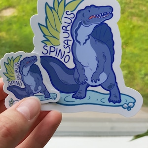 May include: Two blue and green Spinosaurus dinosaur stickers. The larger sticker shows a Spinosaurus standing on a blue surface with a green leafy design behind it. The smaller sticker shows a Spinosaurus standing on a blue surface with a green leafy design behind it.