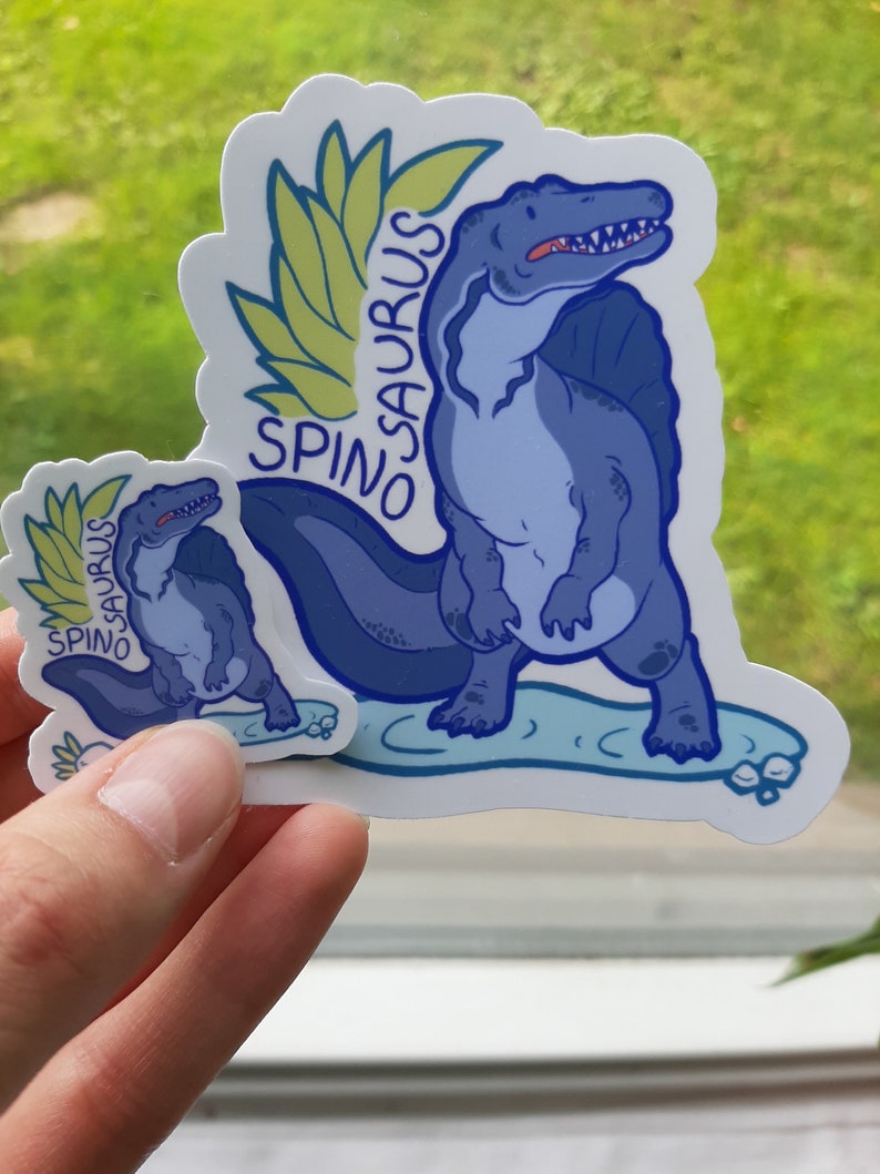 May include: Two blue and green Spinosaurus dinosaur stickers. The larger sticker shows a Spinosaurus standing on a blue surface with a green leafy design behind it. The smaller sticker shows a Spinosaurus standing on a blue surface with a green leafy design behind it.