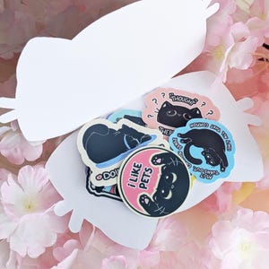 May include: A selection of cat-themed stickers in different shapes and colours. The stickers depict black cat illustrations with phrases such as "I like pets" and "Thought?" on a white cat-shaped card, surrounded by pink flowers.