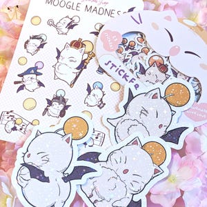 Könnte beinhalten: Eine Sammlung von Aufklebern mit cartoonartigen, katzenartigen Charakteren mit Fledermausflügeln, Kronen und Zaubererhüten. Die Aufkleber sind weiß mit Glitzerakzenten und sind auf einem rosa floralen Hintergrund angeordnet. Der Text "Moogle Madness" ist sichtbar.