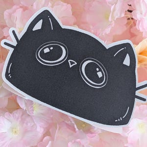 May include: A black cat-shaped sticker with large, round eyes and whiskers. The sticker is set against a backdrop of soft pink and peach-coloured flowers, creating a whimsical and playful aesthetic. The cat's face is simple and cartoonish.