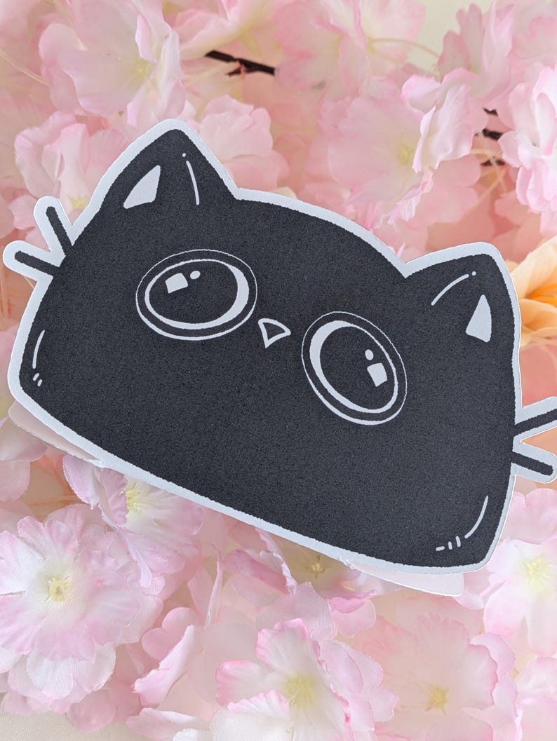 May include: A black cat-shaped sticker with large, round eyes and whiskers. The sticker is set against a backdrop of soft pink and peach-coloured flowers, creating a whimsical and playful aesthetic. The cat's face is simple and cartoonish.