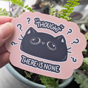 May include: A pink sticker featuring a black cat illustration and the text "THOUGHT THERE IS NONE." The cat has large eyes and question marks surround the cat's head. The sticker is held in front of a background of green plants.
