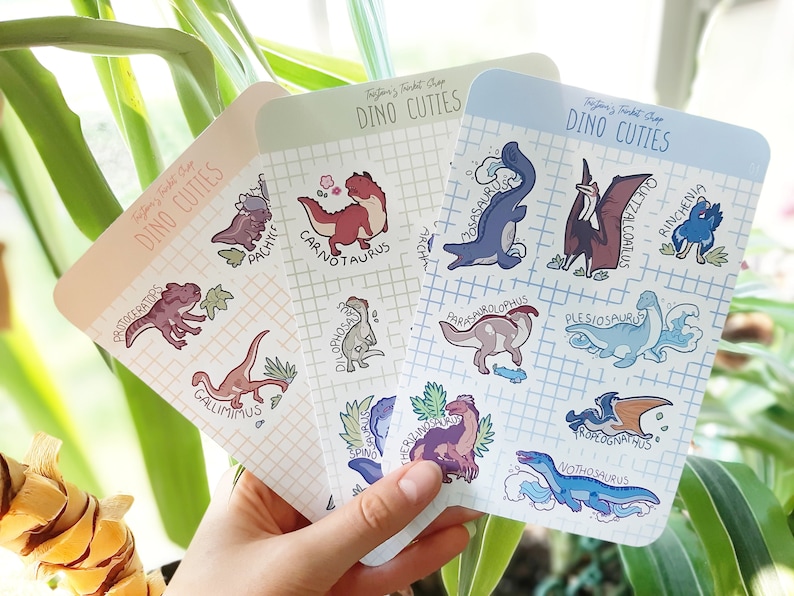 May include: Three cards with a light blue, light green, and peach background. Each card has a grid pattern and features a different set of cartoon dinosaurs with their names written below. The dinosaurs are drawn in a cute style with bright colors. The cards are titled "Dino Cuties".