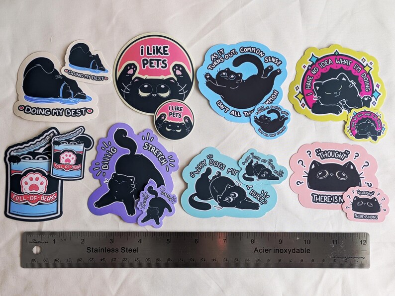 May include: A selection of cat-themed stickers in various shapes and colours. The designs feature black cats with phrases such as "I Like Pets" and "Doing My Best." A stainless steel ruler is included for scale.