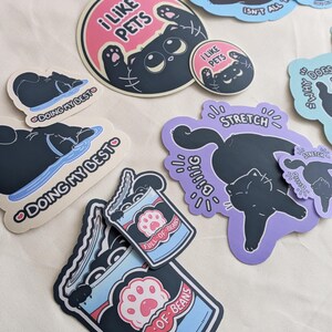 May include: A selection of cat-themed stickers in diverse shapes and sizes. Designs feature a black cat with phrases such as "I Like Pets," "Doing My Best," and "Full of Beans." The stickers have a playful, cartoonish style with pastel colour accents.