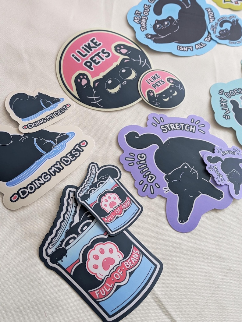 May include: A selection of cat-themed stickers in diverse shapes and sizes. Designs feature a black cat with phrases such as "I Like Pets," "Doing My Best," and "Full of Beans." The stickers have a playful, cartoonish style with pastel colour accents.