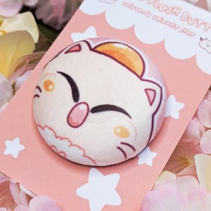 May include: A round plush button featuring a cartoon cat face with a pink nose, orange ears, and a smiling expression. The button is on a pink card with white stars and the text "Moogle Plush Button".