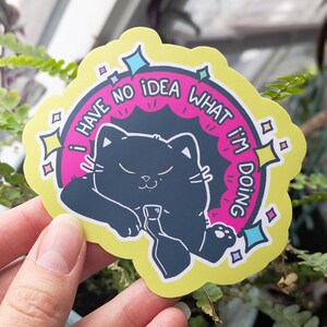 May include: A die-cut sticker featuring a black cat holding a bottle, with the text "I HAVE NO IDEA WHAT I'M DOING" in a pink and yellow design. The sticker has a yellow border with star and diamond accents.