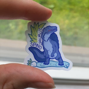 May include: A blue and white sticker featuring a cartoon Spinosaurus dinosaur with a green leafy crown. The text "SPINO SAURUS" is written on the sticker.