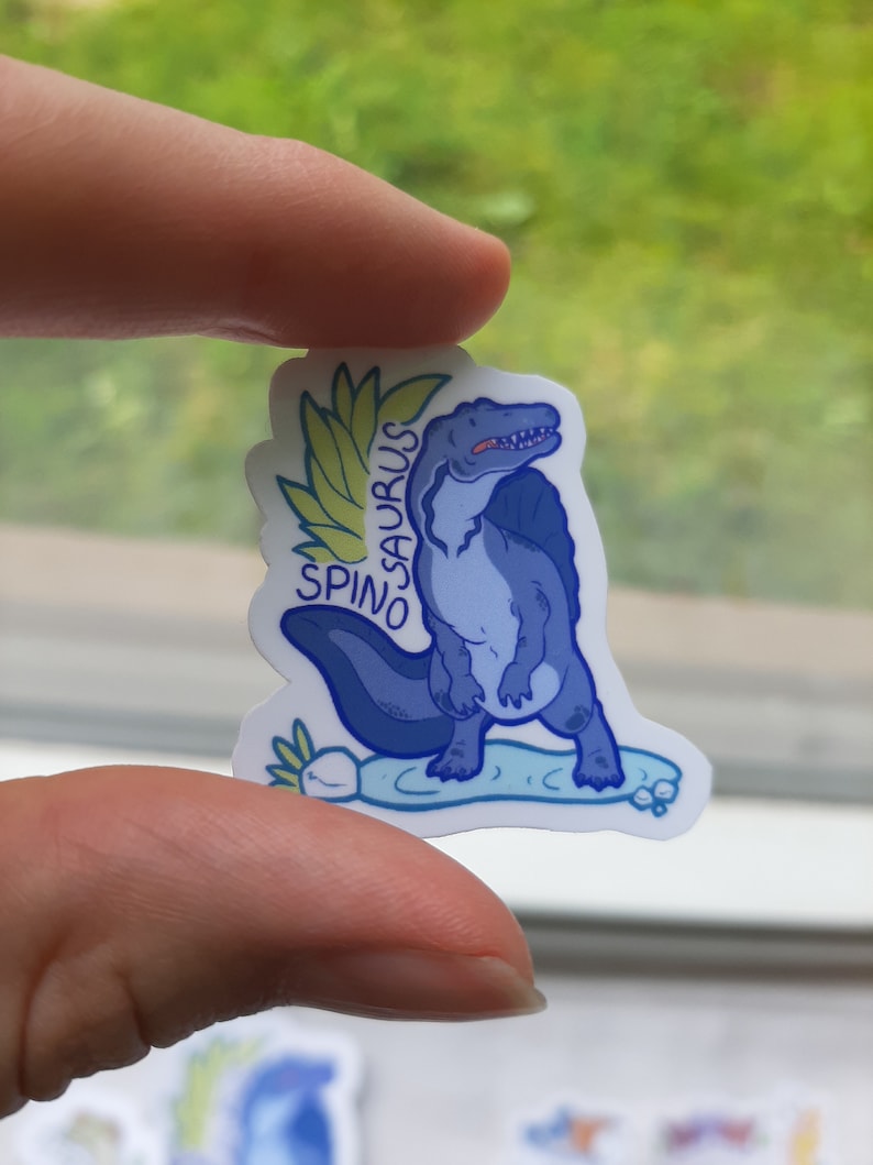 May include: A blue and white sticker featuring a cartoon Spinosaurus dinosaur with a green leafy crown. The text "SPINO SAURUS" is written on the sticker.