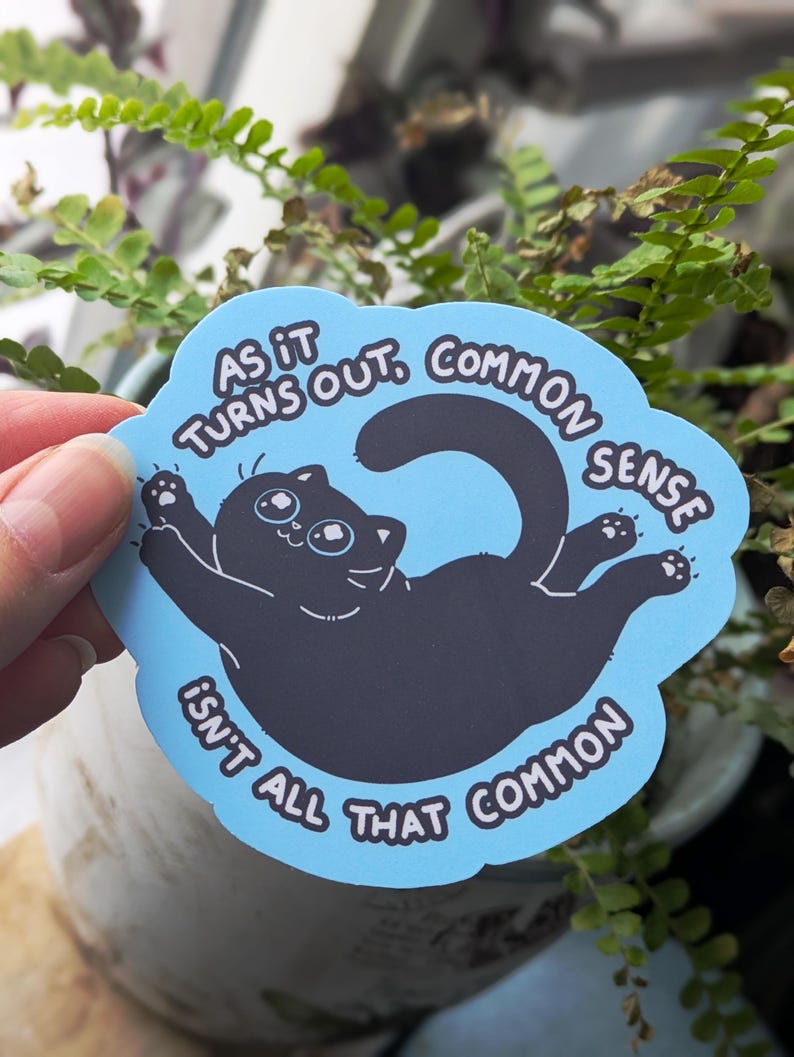 May include: A blue sticker featuring a black cat illustration. The cat is lying on its back with its paws raised. The sticker has the text "AS IT TURNS OUT, COMMON SENSE ISN'T ALL THAT COMMON" in a circular design.