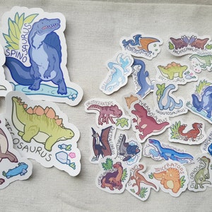 May include: A collection of colorful dinosaur stickers with names like Spinosaurus, Dilophosaurus, and Parasaurolophus. The stickers are on a light beige fabric background.