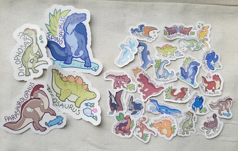May include: A collection of colorful dinosaur stickers with names like Spinosaurus, Dilophosaurus, and Parasaurolophus. The stickers are on a light beige fabric background.