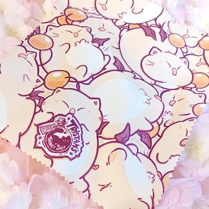 May include: A square fabric featuring a repeating pattern of cartoon cats with small purple wings and orange orbs. The fabric has a logo that says "Cats Trinket Shop" and "Tristans". The background is a soft pink with flowers.
