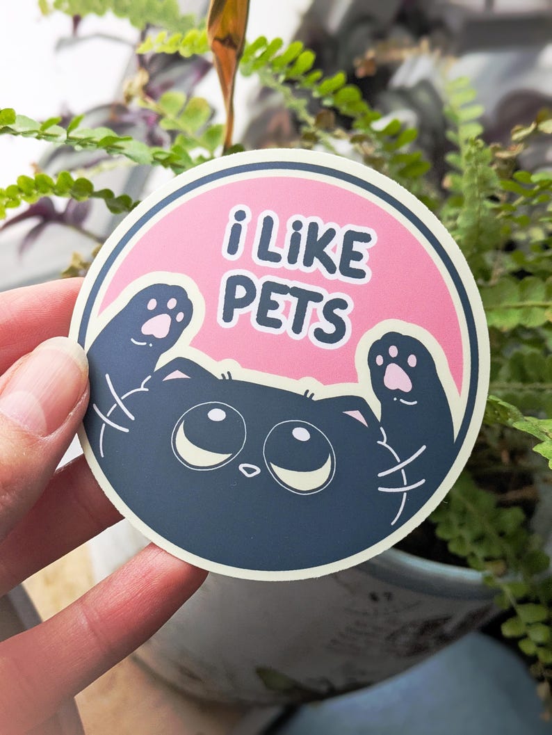 May include: A circular sticker with the words "i LIKE PETS" in white on a pink backdrop. It shows a cartoon cat with big eyes and paws. The sticker is held in front of a plant.