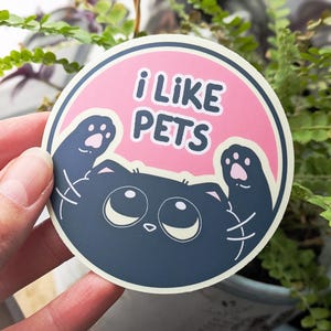 May include: A round sticker with the text "i LIKE PETS" in white on a pink background. The sticker features a cartoon cat with large eyes and paws. The sticker is held in front of a plant.