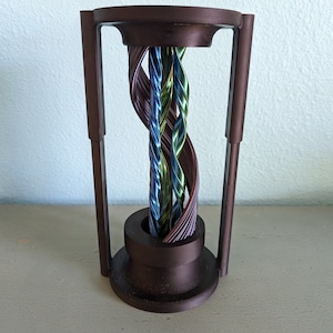 May include: A bronze-colored sculpture with a cylindrical base and top, connected by vertical supports. Inside, twisted strands of blue, green, and purple metallic material create a visually dynamic centerpiece. The sculpture is set against a light-colored background.