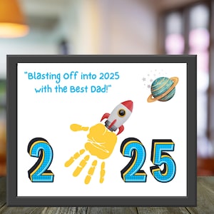 Puede incluir: Impresión enmarcada con temática espacial. La obra presenta un cohete, un planeta y los números 2 y 25. El texto dice "Blasting Off into 2025 with the Best Dad!" Se incorpora una huella de mano amarilla.