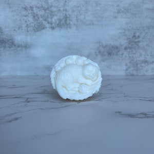Op de afbeelding: Een wit, rond decoratief object met een ontwerp van een slapende kat. De kat is opgerold en het object heeft een getextureerde, wolkenachtige rand. Het object is waarschijnlijk gemaakt van keramiek of een vergelijkbaar materiaal.