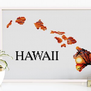 May include: A framed cross-stitch artwork depicting the Hawaiian Islands in shades of orange, red, and yellow, with the word "HAWAII" in bold black letters. The artwork is set against a white background.