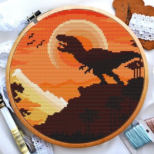 Könnte beinhalten: Nahaufnahme eines Kreuzstich-Stickrahmens mit einer Sonnenuntergangsszene, die eine Silhouette eines Dinosauriers vor einem großen, orangefarbenen Mond zeigt. Der Hintergrund ist ein Farbverlauf von Orange, Gelb und Braun.