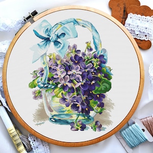 May include: A cross-stitch embroidery of a basket overflowing with purple violets, tied with a light blue bow. The embroidery is set in a wooden hoop, with spools of thread and lace trim in the background.