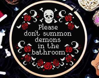 May include: A black cross-stitch hoop with the text "Please don't summon demons in the bathroom." The design includes a skull, crossbones, and red roses. The embroidery is set in a wooden hoop.
