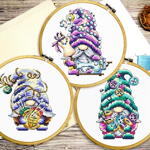 May include: Three cross-stitch embroidery hoops with colorful gnome designs. The gnomes are wearing hats decorated with buttons, yarn, and beads. Each gnome is holding a sewing-related item, such as a spool of thread, a sewing machine, or a needle and thread.