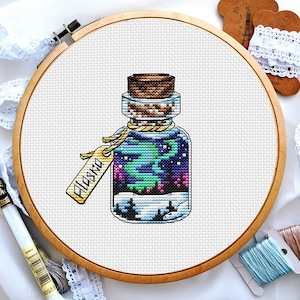 May include: A cross-stitch pattern of a glass bottle with a cork stopper. The bottle contains a swirling blue, green, and purple liquid with a white label that says "Alaska".
