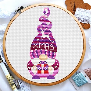 May include: A cross-stitch pattern of a purple and pink gnome wearing a hat that says "XMAS" with a reindeer design. The gnome is holding a gift box.