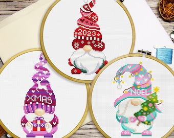 Può includere: Tre progetti di punto croce in cerchi di legno. Ogni disegno presenta uno gnomo con un cappello alto. Uno ha un cappello rosso con "2023", un altro un cappello viola con "XMAS" e il terzo un cappello turchese con "NOEL".