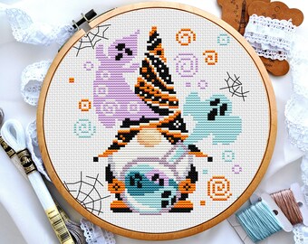 Op de afbeelding: Een kruissteekpatroon van een Halloween-dwerg die een heksenhoed draagt met oranje en zwarte strepen. De dwerg houdt een pompoen vast met een griezelig gezicht. De achtergrond is wit met paarse en blauwe geesten, oranje en zwarte spinnenwebben en oranje cirkels.
