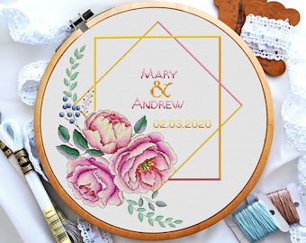 Puede incluir: Un aro de bordado blanco con un diseño de punto de cruz que presenta un ramo de flores, un marco geométrico dorado y rosa, y el texto "Mary & Andrew 02.03.2020".