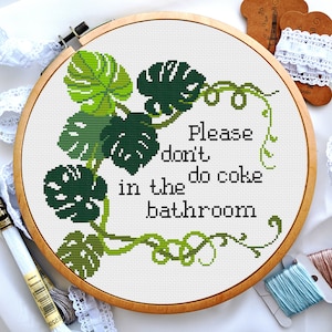 May include: A white embroidery hoop with a green and white cross-stitch design. The design features a vine with monstera leaves and the text "Please don't do coke in the bathroom".
