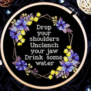 Op de afbeelding: Borduurwerk op een borduurring met een ontwerp van een bloemkrans. De tekst luidt "Laat je schouders zakken Ontspan je kaak Drink wat water".