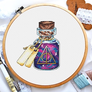 May include: A cross-stitch pattern of a glass bottle filled with a swirling purple and pink liquid. The bottle has a cork stopper and a gold string tied around the neck. The liquid has a gold triangle symbol inside.