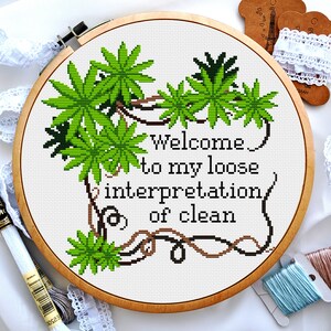 Könnte beinhalten: Ein Kreuzstich-Stickrahmen mit einem grünen und braunen Design aus Ranken und Blättern. Der Text "Welcome to my loose interpretation of clean" ist in weißer Farbe gestickt.