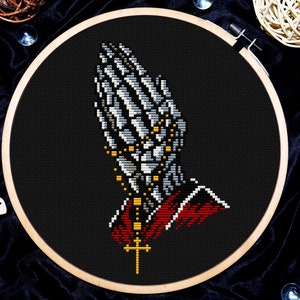 May include: Cross stitch embroidery hoop with a black background and a white skeleton hand in a praying position with a gold rosary and a red and gold robe.