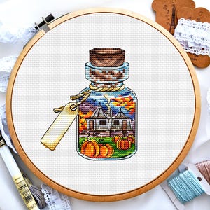 May include: A cross-stitch pattern of a glass jar with a cork stopper filled with a miniature house and pumpkins. The jar is on a white background.
