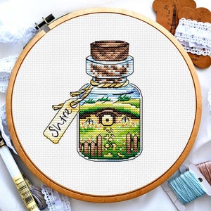 May include: A cross-stitch embroidery hoop with a colorful image of a glass bottle with a cork stopper. The bottle contains a miniature house and a green field with flowers. The bottle has a tag that says "Shine".
