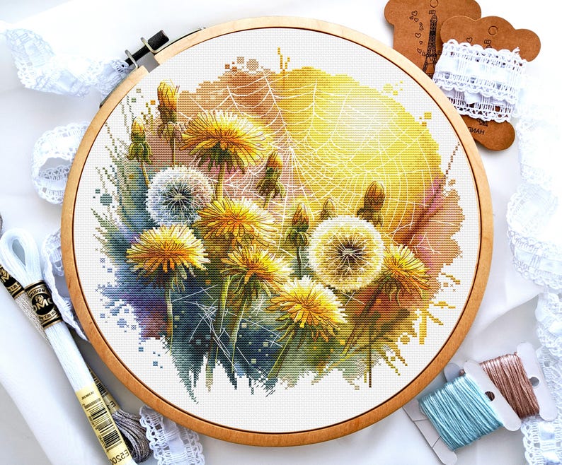 May include: A cross-stitch embroidery of dandelions in a wooden hoop. The design features yellow and white dandelions with a watercolor effect, set against a yellow and orange background. Embroidery floss and lace are visible.