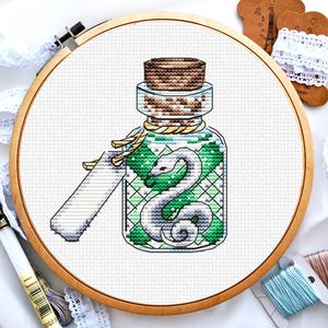 Puede incluir: Un patrón de punto de cruz de una botella de vidrio con un tapón de corcho, que contiene un líquido verde y una serpiente blanca. La botella tiene una etiqueta que dice "Basilisk"