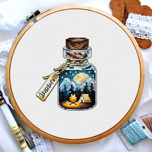 May include: A cross-stitch embroidery of a glass bottle with a cork stopper. The bottle contains a snowy mountain scene with a campfire and a tent. The bottle has a tag that says "Adventure".