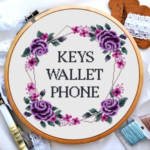 Peut inclure: Broderie au point de croix dans un cercle en bois. Le motif présente une couronne florale avec des roses violettes et des fleurs roses, entourant les mots "KEYS WALLET PHONE" en noir. Fils à broder et dentelle visibles.