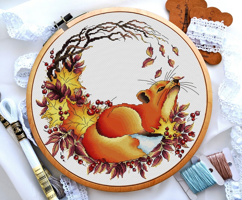 Op de afbeelding: Een kruissteekpatroon van een vos die opgerold ligt in een cirkel van herfstbladeren. De vos is oranje en wit, en de bladeren zijn rood, oranje en geel. Het patroon is ingelijst in een houten ring.