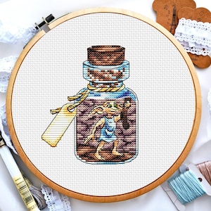 May include: A cross-stitch pattern of a brown glass bottle with a cork stopper. Inside the bottle is a small, brown, winged creature with large ears and blue eyes. The creature is holding a small, brown object in its hand.