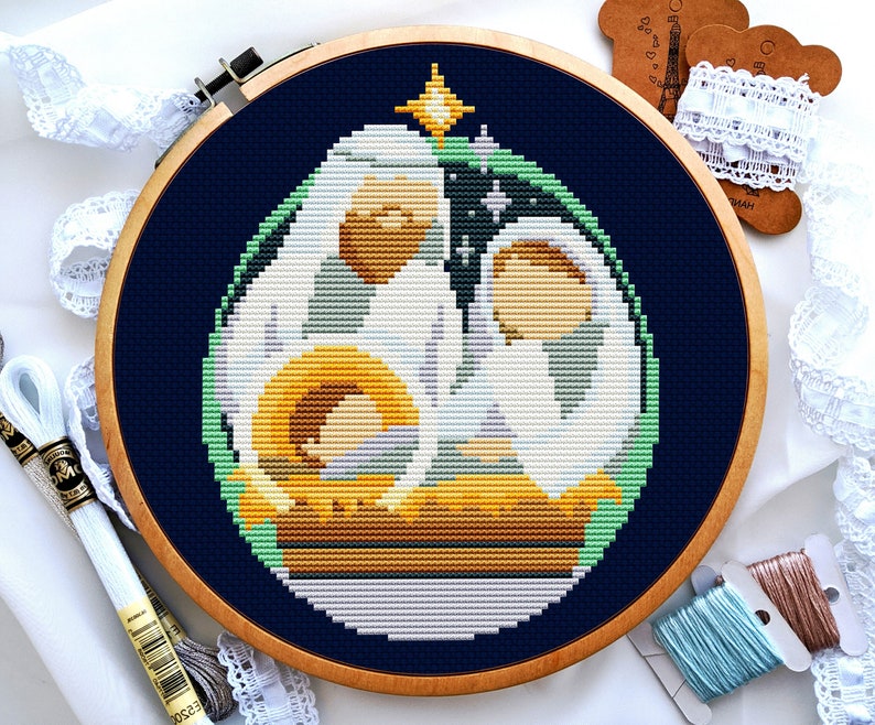 May include: A cross-stitch embroidery of a nativity scene in a wooden hoop. The design features Mary, Joseph, and baby Jesus in a stable, with a star above. The background is navy blue, and the embroidery uses white, yellow, and green thread.