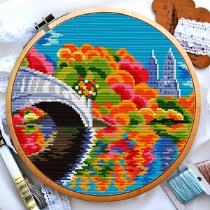 May include: A colourful cross-stitch embroidery of a bridge over a river with buildings in the background. The embroidery is framed in a wooden hoop.